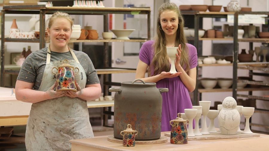 Two Ceramics Program students stand in the shop with their works that are in progress.