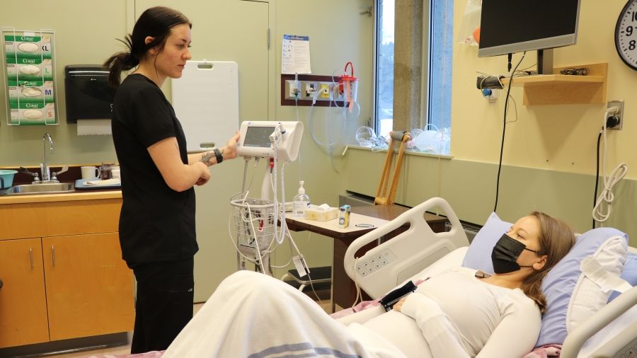 A nursing student stands at the bedside of a mock patient in a hospital bed